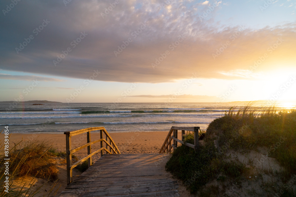 A wooden bridge leads to the beach. The sky is cloudy and the sun is setting. The beach is calm and peaceful. A wooden bridge leads to the beach. The sky is cloudy and the sun is setting. The beach is calm and peaceful.