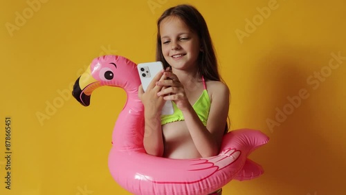 Smiling cheerful little girl in flamingo rubber ring around waist isolated over yellow background scrolling online browsing web pages playing kid's games