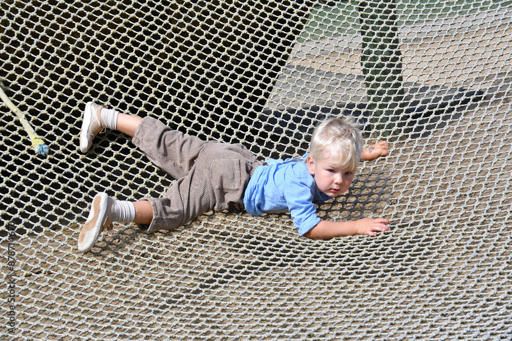 Naklejka premium Happy little boy climbing on the rope net in adventure activity park
