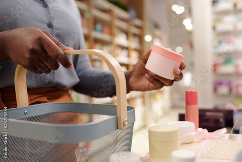 Unrecognizable female consumer with shopping basket holding jar of facial cream while standing by display with cosmetic products photo