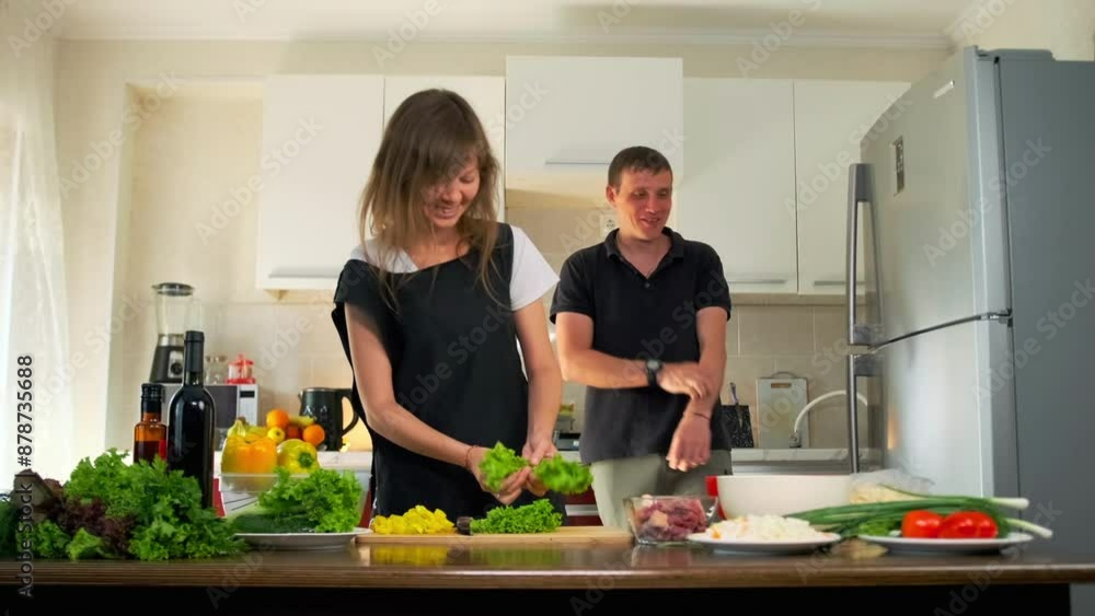 Happy married couple preparing dinner together, young wife smiling cleaning salad leaves standing in the kitchen with her husband near fresh vegetables. Happy married couple preparing dinner together, young wife smiling cleaning salad leaves standing in the kitchen with her husband near fresh vegetables.