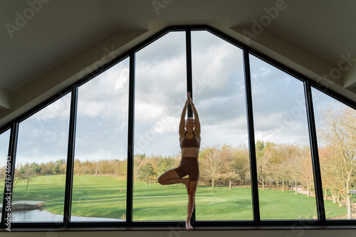 Silhouette of a woman practicing yoga vinyasa flow, tree balancing pose indoors next to the window. Wellness, lifestyle, horizontal, copy space.