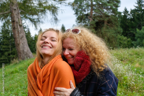 Two smiling women wrapped in warm blankets, embracing each other in a lush, green forest. The younger woman beams with happiness while the older woman exudes warmth and love