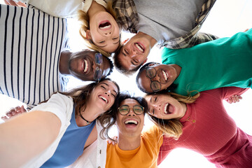 Selfie from below of large group of diverse friends posing hugging in circle looking excited at camera. Cheerful young people standing embracing outdoor. Photo of generation z enjoying sunny meeting