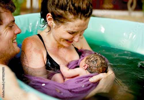 Water birth. Home birth. A woman cradles a newborn baby wrapped in a purple towel while sitting in a birth pool. Her husband is next to her, smiling and sharing in the joyful moment.