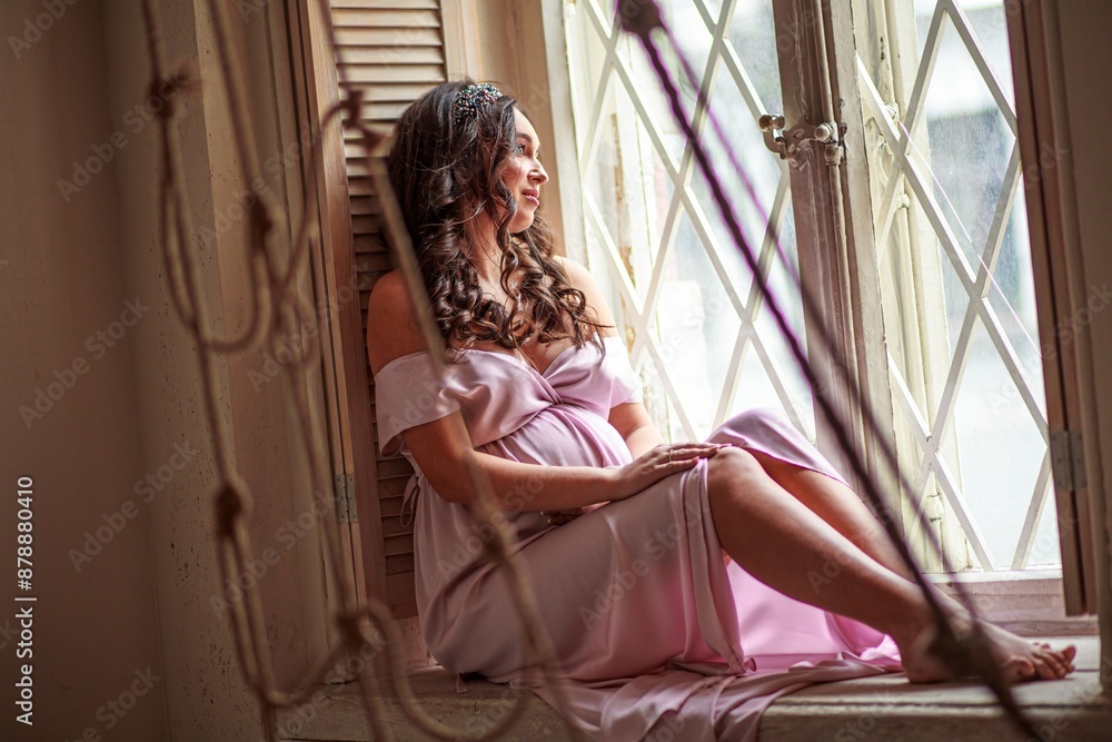 Beautiful pregnant woman sitting on the windowsill and looking out the window Beautiful pregnant woman sitting on the windowsill and looking out the window