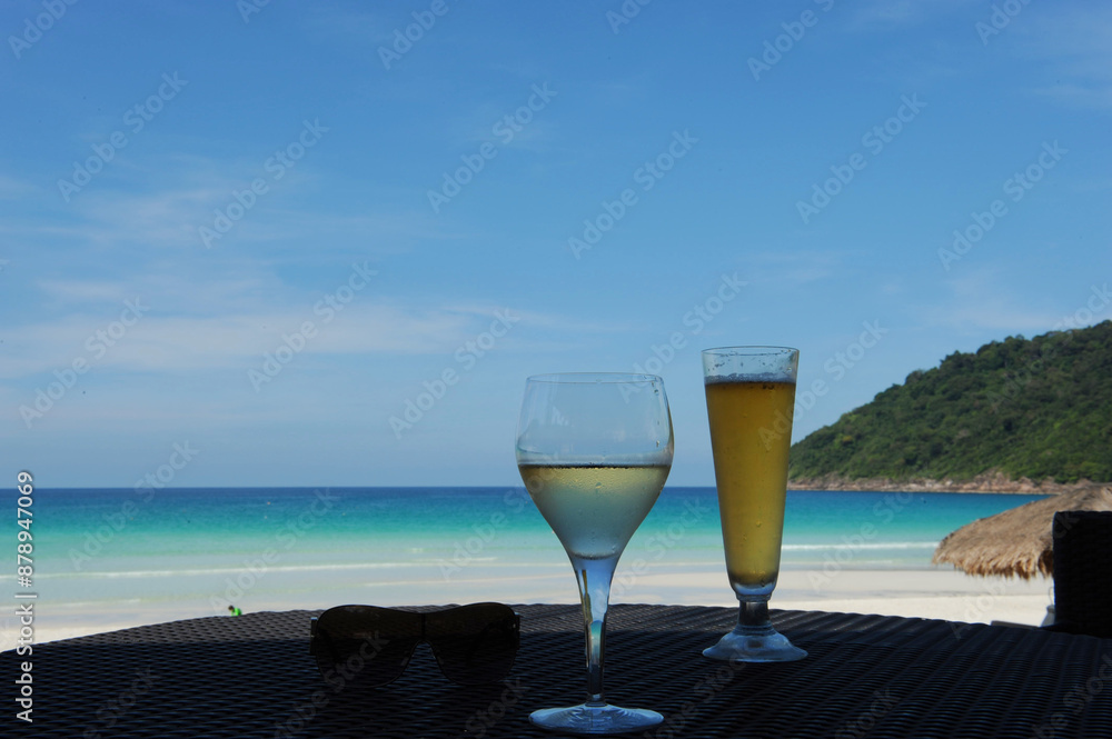 Two glasses of drinks, one is beer and the other is white wine against a scenic white sandy beach, a mountain and blue sky background. Two glasses of drinks, one is beer and the other is white wine against a scenic white sandy beach, a mountain and blue sky background.