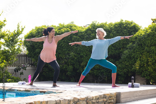 Two happy diverse senior female friends practicing yoga lunges stretching in sunny garden