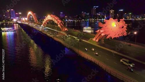 Dragon Bridge in Da Nang, Vietnam, beautifully illuminated at night. The scene captures various vehicles crossing the bridge, while boats navigate the river below.