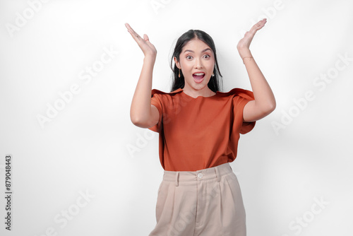 Excited young Asian woman in a brown shirt and taupe pants is pointing to the copy space above her, isolated by white background. photo