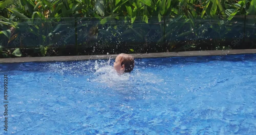 Joyful toddler stands in shallow wading pool, splashing water with his hands and jumping up and down, creating large splashes. On sunny, hot day, baby boy energetically plays in water Joyful toddler stands in shallow wading pool, splashing water with his hands and jumping up and down, creating large splashes. On sunny, hot day, baby boy energetically plays in water