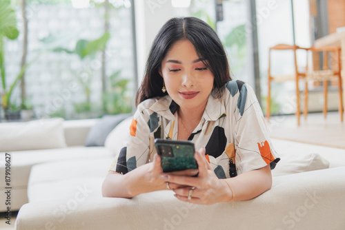 Peaceful young Asian woman is sitting on a couch and looking at her cell phone. She is smiling and enjoying free time in social media. #879108627