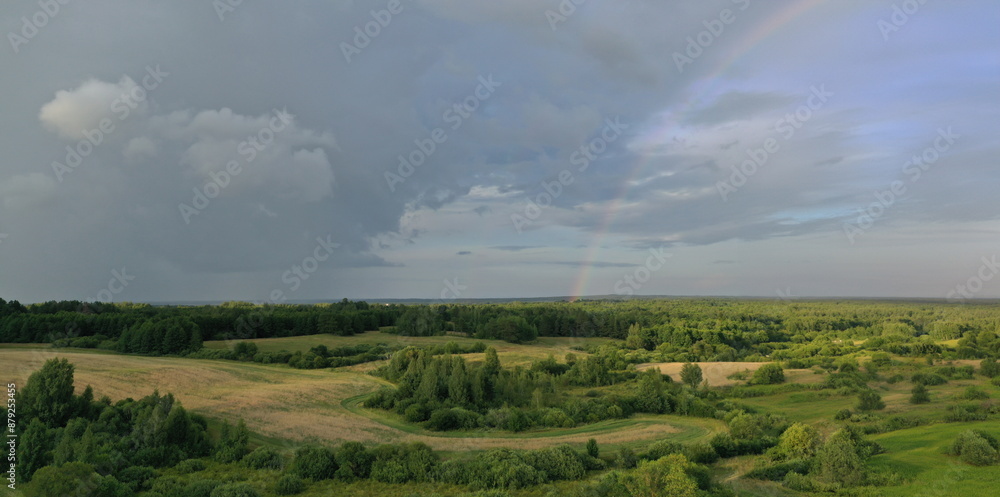 Fototapeta premium Top view of meadow with road and green plants. Cloudy sky and beautiful rainbow above trees and bushes. Picturesque landscape scene of rain in summer time