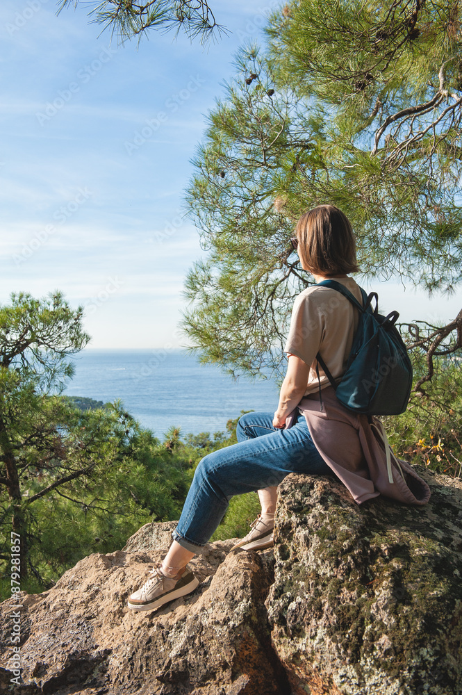Naklejka premium Woman with a backpack looking out at a scenic view of hills and trees, sitting on a rock