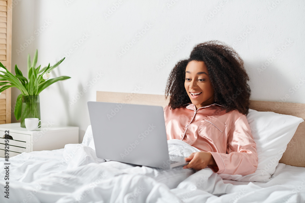 African American woman in pajamas sitting on bed, using laptop in bedroom in the morning. African American woman in pajamas sitting on bed, using laptop in bedroom in the morning.