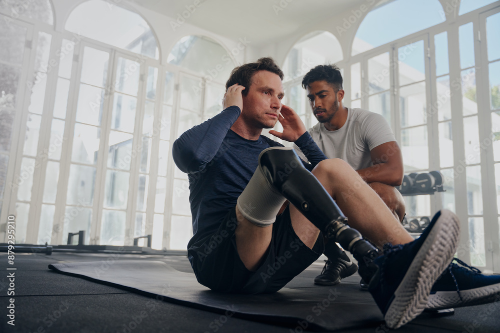 Obraz premium Multiracial trainer next to young man in sportswear with prosthetic leg doing push-ups at the gym