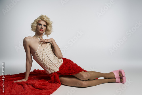 Blonde drag queen in red skirt and corset, legs stretched out, against grey backdrop photo