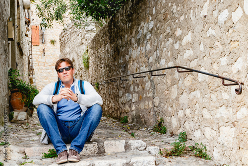 A traveler in sunglasses and a backpack relaxes in a peaceful alley with rustic architecture. Saint Paul de Vence. France. Provence. photo