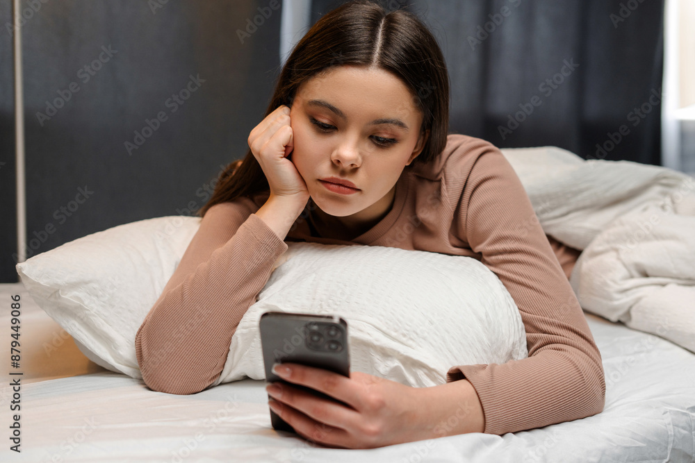 Young, sad girl, teenager lying on bed is feeling sad while using smartphone Young, sad girl, teenager lying on bed is feeling sad while using smartphone