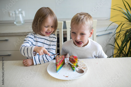 The older brother and little sister are laughing and eating a multicolored cake.