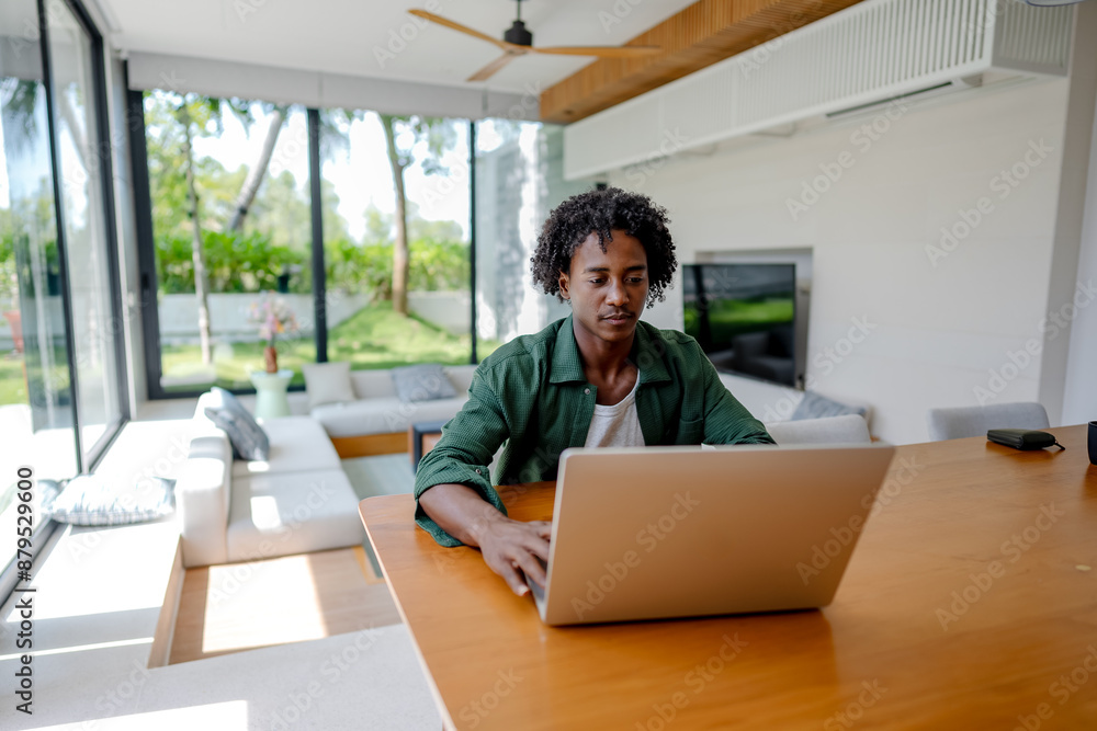 A young man of African descent sitting in a modern living room, looking at the laptop. His expression is one of concentration or concern, he is solving a problem or thinking about home finances A young man of African descent sitting in a modern living room, looking at the laptop. His expression is one of concentration or concern, he is solving a problem or thinking about home finances