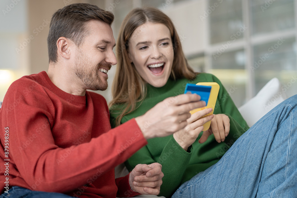 Joyful happy young family couple making secure internet payment in smartphone app sits at home. Contented spouses man and woman using phone shopping online, paying by credit card, looking at screen. Joyful happy young family couple making secure internet payment in smartphone app sits at home. Contented spouses man and woman using phone shopping online, paying by credit card, looking at screen.