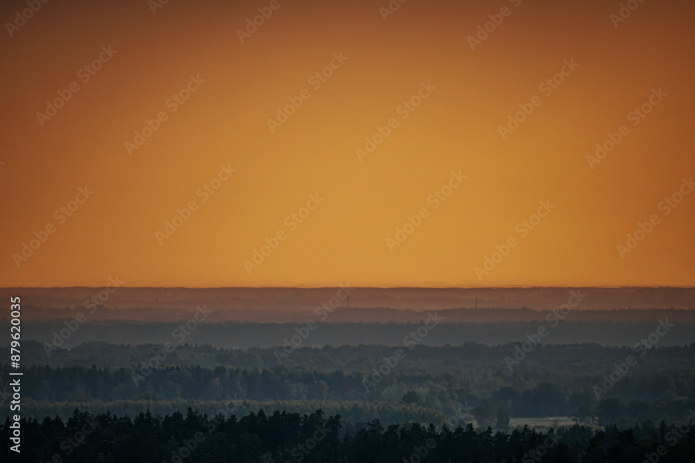 Obraz premium Wind turbines stand in the distance as the sun sets over a dense forest landscape. The sky glows with warm orange hues, creating a serene and atmospheric scene.