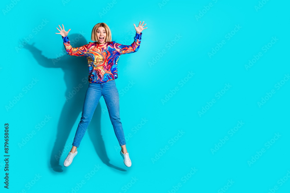 Full size photo of astonished ecstatic girl dressed print shirt jeans jumping near empty space arms up isolated on blue color background Full size photo of astonished ecstatic girl dressed print shirt jeans jumping near empty space arms up isolated on blue color background