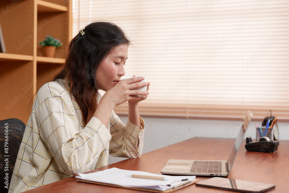 Asian female office worker taking a sip from a cup while seated at a wooden desk. Casual attire in a modern office setting with documents and a tablet on the desk. Relaxed and thoughtful expression. Asian female office worker taking a sip from a cup while seated at a wooden desk. Casual attire in a modern office setting with documents and a tablet on the desk. Relaxed and thoughtful expression.