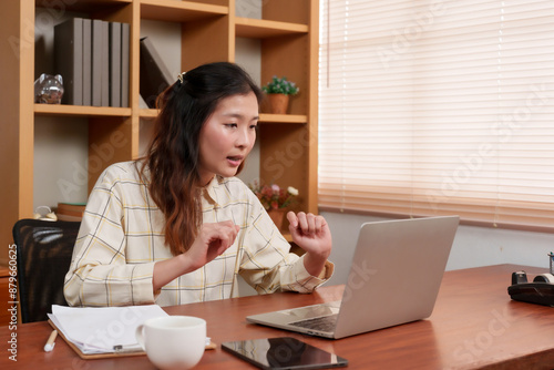 Beautiful Asian female office worker engaging in a video call on a laptop. Animated expression with hands raised. Seated at a desk in a modern office with wooden shelves in the background.