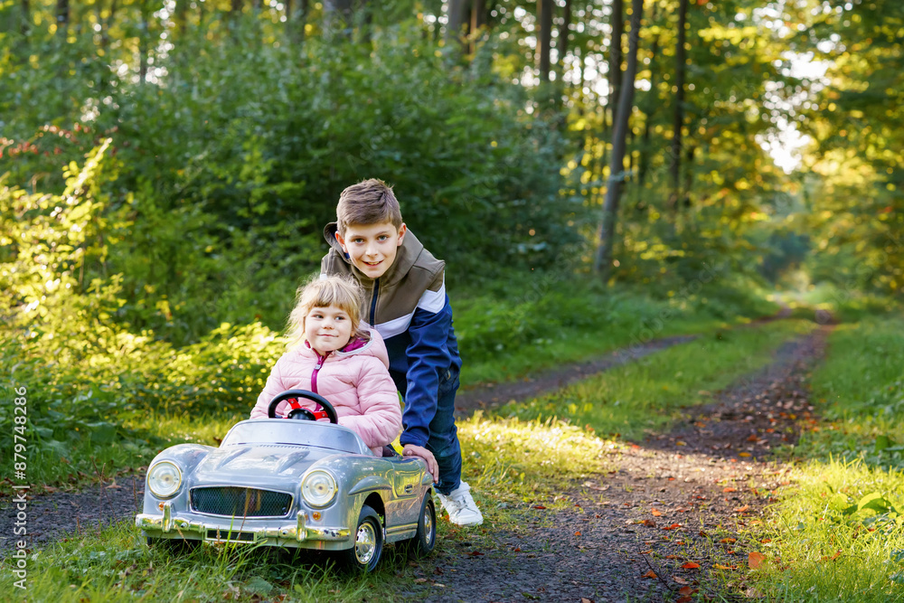 Two happy children playing with big old toy car in autumn forest, outdoors. Kid boy pushing and driving car with little toddler girl, cute sister inside. Laughing and smiling kids. Lovely family Two happy children playing with big old toy car in autumn forest, outdoors. Kid boy pushing and driving car with little toddler girl, cute sister inside. Laughing and smiling kids. Lovely family