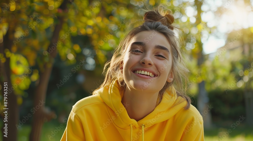 A young woman in a yellow hoodie smiles warmly in an outdoor setting filled with greenery and trees, enjoying the pleasant weather and nature around her. A young woman in a yellow hoodie smiles warmly in an outdoor setting filled with greenery and trees, enjoying the pleasant weather and nature around her.