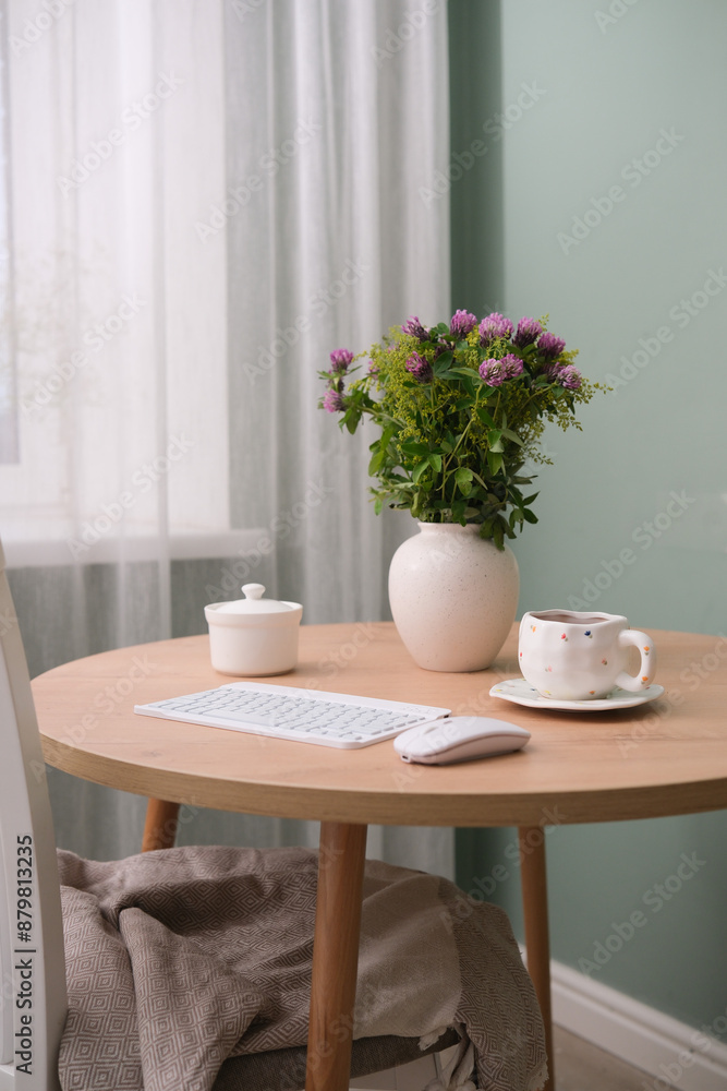 Home workplace in interior - keyboard, computer mouse, cup of coffee, vaze with flowers on wooden table near the window in living room. Home workplace in interior - keyboard, computer mouse, cup of coffee, vaze with flowers on wooden table near the window in living room.