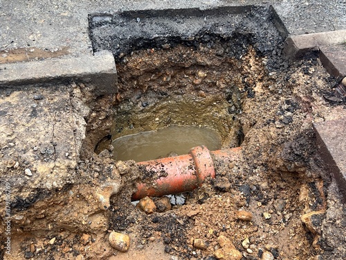 Close up of broken water pipe in construction site in the ground showing drains dug up on road with water works with earth rubble flooding bricks cement stone and soil in layers in urban city street