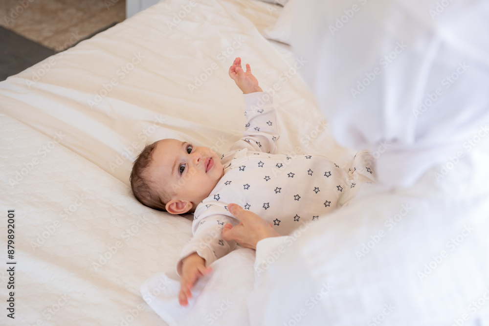 Baby in star-patterned pajamas lying on a white bed with a teddy bear, attended by a parent in hijab. Top view, closeup, perfect for family and parenting themes Baby in star-patterned pajamas lying on a white bed with a teddy bear, attended by a parent in hijab. Top view, closeup, perfect for family and parenting themes