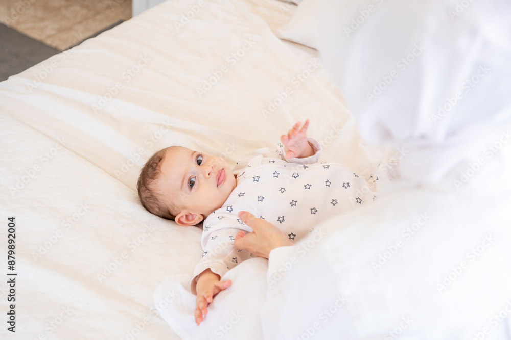 Baby in star-patterned pajamas lying on a white bed with a teddy bear, attended by a parent in hijab. Top view, closeup, perfect for family and parenting themes Baby in star-patterned pajamas lying on a white bed with a teddy bear, attended by a parent in hijab. Top view, closeup, perfect for family and parenting themes