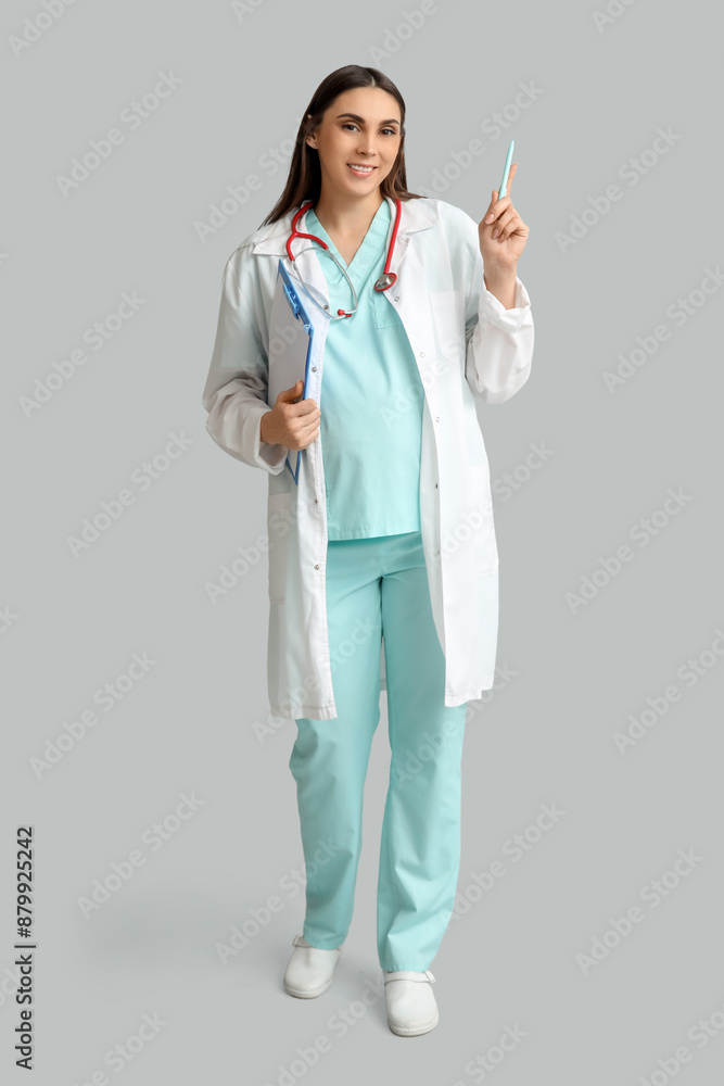Beautiful young pregnant doctor holding clipboard on grey background Beautiful young pregnant doctor holding clipboard on grey background