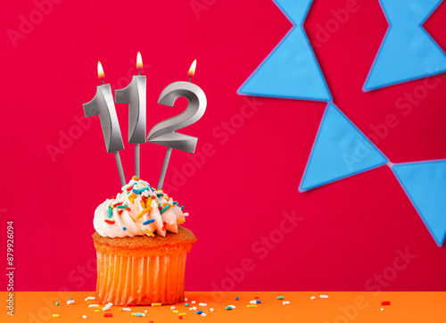 Number 112 candle with birthday cupcake on a red background with blue pennants