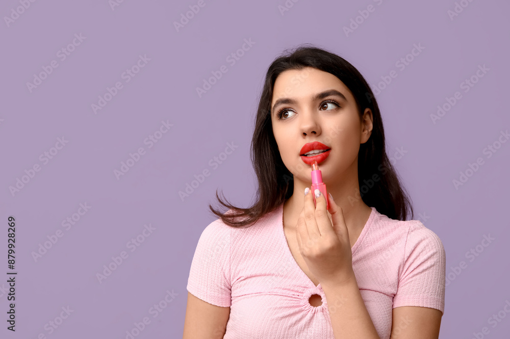 Fototapeta premium Beautiful young woman applying lipstick on lilac background