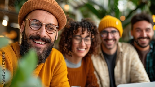 Four friends wearing glasses and warm clothing, two with beanies, smiling and sitting close together in a cozy cafe with green plants, enjoying their bonding time.