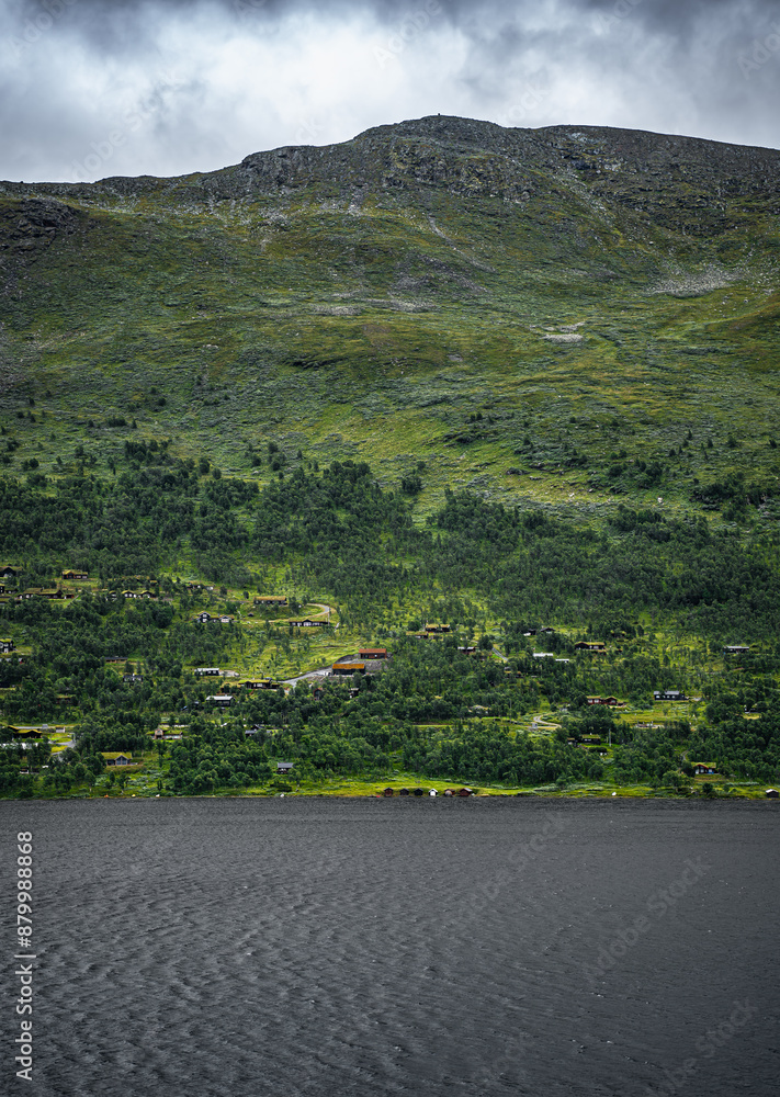 Fototapeta premium Holiday homes sit by a lake surrounded by green, beneath the steep mountain range. Storm clouds gather above the rocky outcrop, contrasting with the serene scene