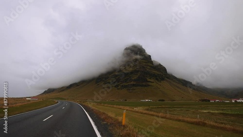 Driving down a highway in Iceland next to a mountain covered by low clouds and fog.