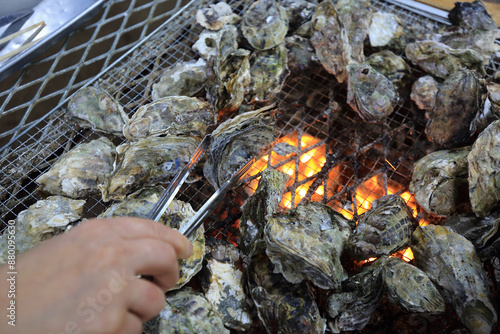 Quadro su tela Close-up of a man's hand and tongs holding an oyster being roasted on the grill