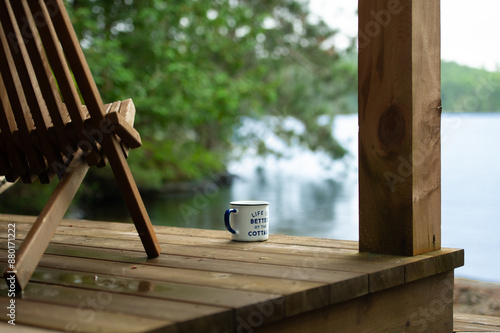 A coffee mug with the phrase "Life is better at the Cottage" rests beside a chair on the wooden porch of a boathouse, overlooking the serene blue waters of a lake in Muskoka, Ontario, Canada.