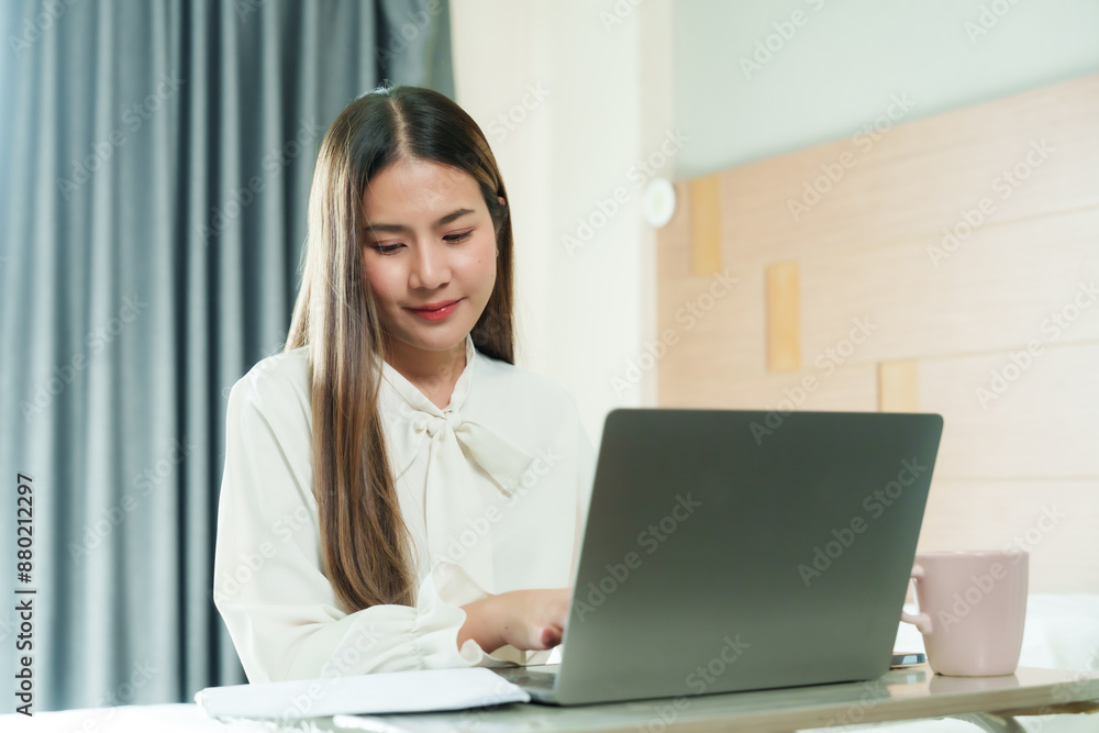 Beautiful Asian woman working from home on holiday, smiling while using laptop, pink cup on table. Cozy bedroom setting, modern lifestyle, balancing work and relaxation, enjoying remote work benefits. Beautiful Asian woman working from home on holiday, smiling while using laptop, pink cup on table. Cozy bedroom setting, modern lifestyle, balancing work and relaxation, enjoying remote work benefits.