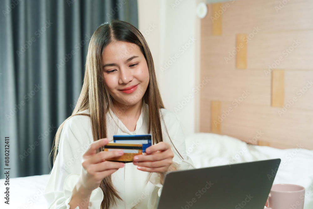 Asian woman working at home, focusing on credit card and laptop for online purchases, cozy bedroom setting. Modern lifestyle, showcasing convenience of remote work and online shopping. Asian woman working at home, focusing on credit card and laptop for online purchases, cozy bedroom setting. Modern lifestyle, showcasing convenience of remote work and online shopping.