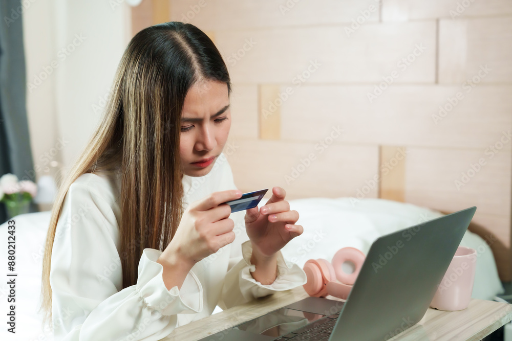 Asian woman working at home, examining credit card details, laptop and pink cup on table. Cozy bedroom setting, modern lifestyle, illustrating remote work and careful online shopping practices. Asian woman working at home, examining credit card details, laptop and pink cup on table. Cozy bedroom setting, modern lifestyle, illustrating remote work and careful online shopping practices.
