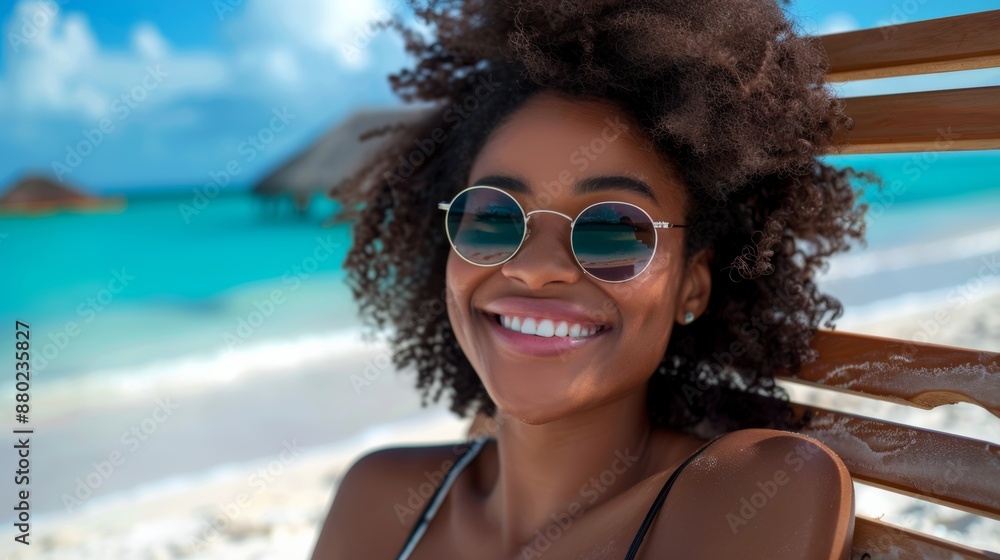 Happy young black woman relaxing on a wooden chair at a tropical beach while looking at the camera wearing glasses. Smiling African American girl with fashion sunglasses enjoying vacation.