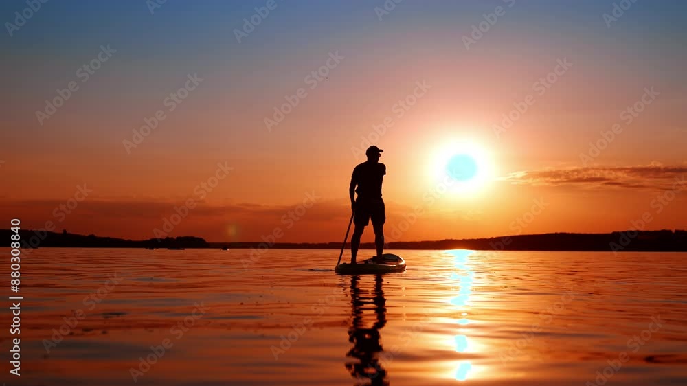Male silhouette distancing from camera on the paddle board. Man oars standing on the inflatable sup board floating by the river at sunset. Low angle view. Male silhouette distancing from camera on the paddle board. Man oars standing on the inflatable sup board floating by the river at sunset. Low angle view.
