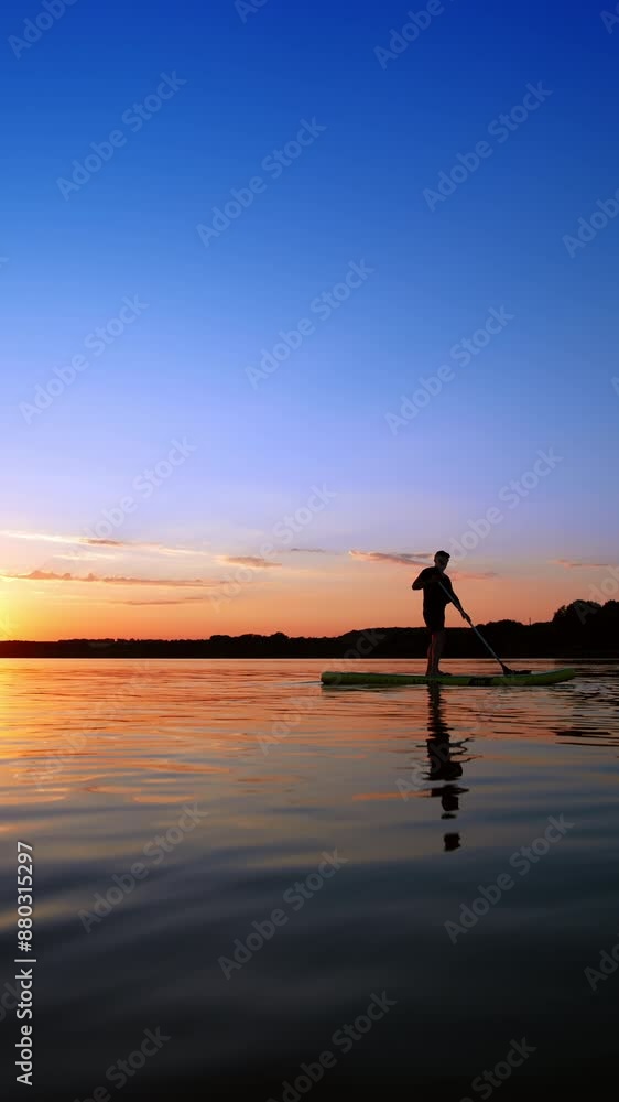 Sportsman practices paddle boarding on the river at sunset. Man stands on the sup board moving with an oar. Vertical video. Sportsman practices paddle boarding on the river at sunset. Man stands on the sup board moving with an oar. Vertical video.